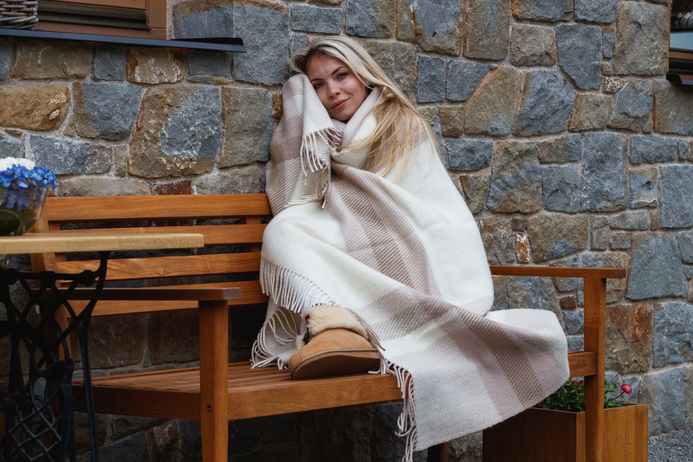 Žena zabalená do světlého vlněného plédu s třásněmi na lavici u kamenné zdi